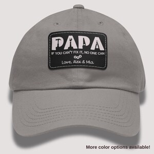 May include: A gray baseball cap with a black patch that reads "PAPA" above the text "IF YOU CAN'T FIX IT, NO ONE CAN" and "Love, Alex & Mia". The patch features tool icons. The hat has a curved brim.