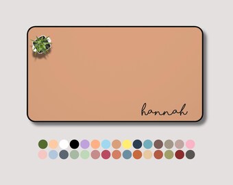May include: A rectangular desk mat in a warm peach color with a black border. The mat features the name "hannah" in a handwritten font and a small potted plant. A color palette of various shades is displayed below.