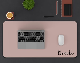 May include: A top-down view of a desk setup featuring a pink desk mat with the name "Brooke" in script. A laptop, white mouse, pen, pencil, notebook, and a cup of coffee are arranged on the desk.