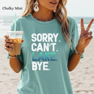 May include: Chalky mint green t-shirt with the text "SORRY. CAN'T. LAKE. BYE." in white and blue. The shirt is a solid color and has short sleeves. The image shows a person wearing the shirt.