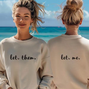May include: Two people wearing beige sweatshirts with the text "let. them." and "let. me." printed on the front and back respectively. The people are standing in front of a beach scene.