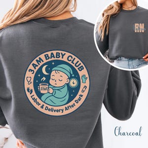May include: Charcoal gray sweatshirt featuring a circular graphic on the back with the text "3AM BABY CLUB" and a sleeping baby illustration. The front of the sweatshirt has a small design with the letters "RN" and the name "Valerie".