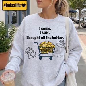 May include: A light gray crewneck sweatshirt featuring the black text I came. I saw. I bought all the butter. The graphic includes a stand mixer, a shopping cart filled with yellow butter blocks, a tiered cake, and a piping bag illustration.