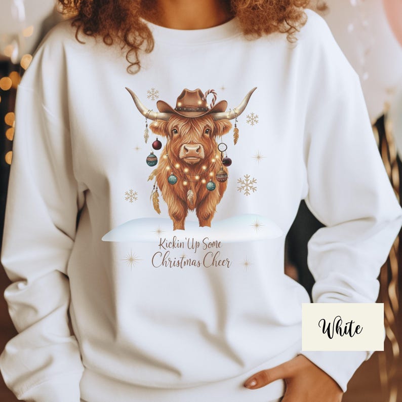 Puede incluir: Sudadera blanca con una vaca de las Tierras Altas marr&oacute;n que lleva un sombrero de vaquero y adornos navide&ntilde;os. La vaca est&aacute; adornada con luces de cadena y plumas. El texto "Kickin' Up Some Christmas Cheer" est&aacute; debajo.