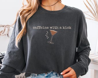 Op de afbeelding: Donkergrijze longsleeve met de tekst "caffeine with a kick" en een cocktailglasafbeelding. Het glas bevat een bruine vloeistof en heeft witte stralende lijnen.