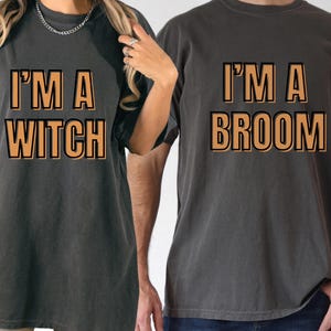 May include: Two dark gray t-shirts with the phrases "I'M A WITCH" and "I'M A BROOM" printed in orange block letters with a black outline. The shirts are likely part of a matching set or couple's costume.