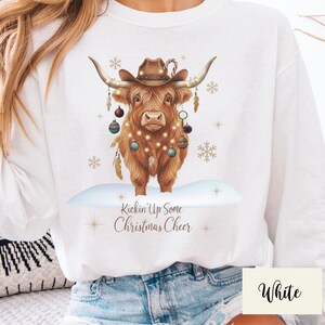 Puede incluir: Sudadera blanca con una vaca de las Tierras Altas marr&oacute;n que lleva un sombrero de vaquero y adornos navide&ntilde;os. La vaca est&aacute; decorada con luces de hadas y el texto "Kickin' Up Some Christmas Cheer" est&aacute; debajo.