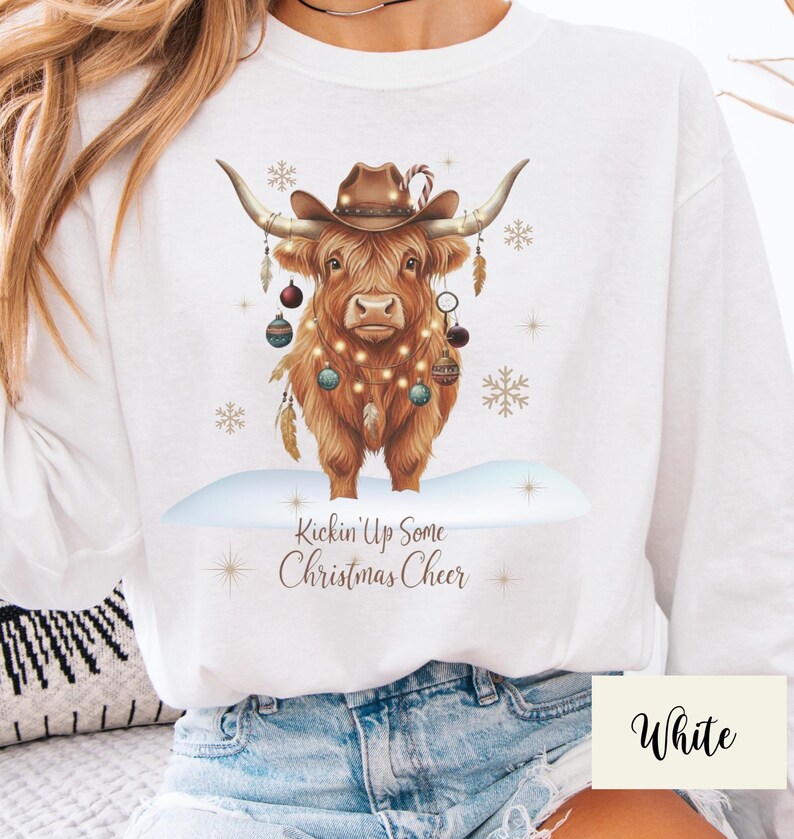 Puede incluir: Sudadera blanca con una vaca de las Tierras Altas marr&oacute;n que lleva un sombrero de vaquero y adornos navide&ntilde;os. La vaca est&aacute; decorada con luces de hadas y el texto "Kickin' Up Some Christmas Cheer" est&aacute; debajo.