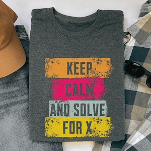 May include: Dark gray t-shirt with the text "KEEP CALM AND SOLVE FOR X" in orange, pink, light blue, and yellow distressed block letters. The shirt is folded and surrounded by other clothing items.