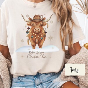 Puede incluir: Camiseta color marfil con una ilustraci&oacute;n festiva de una vaca de las Tierras Altas. La vaca lleva un sombrero de vaquero y est&aacute; adornada con adornos y luces navide&ntilde;as. El texto "Kickin' Up Some Christmas Cheer" est&aacute; debajo.
