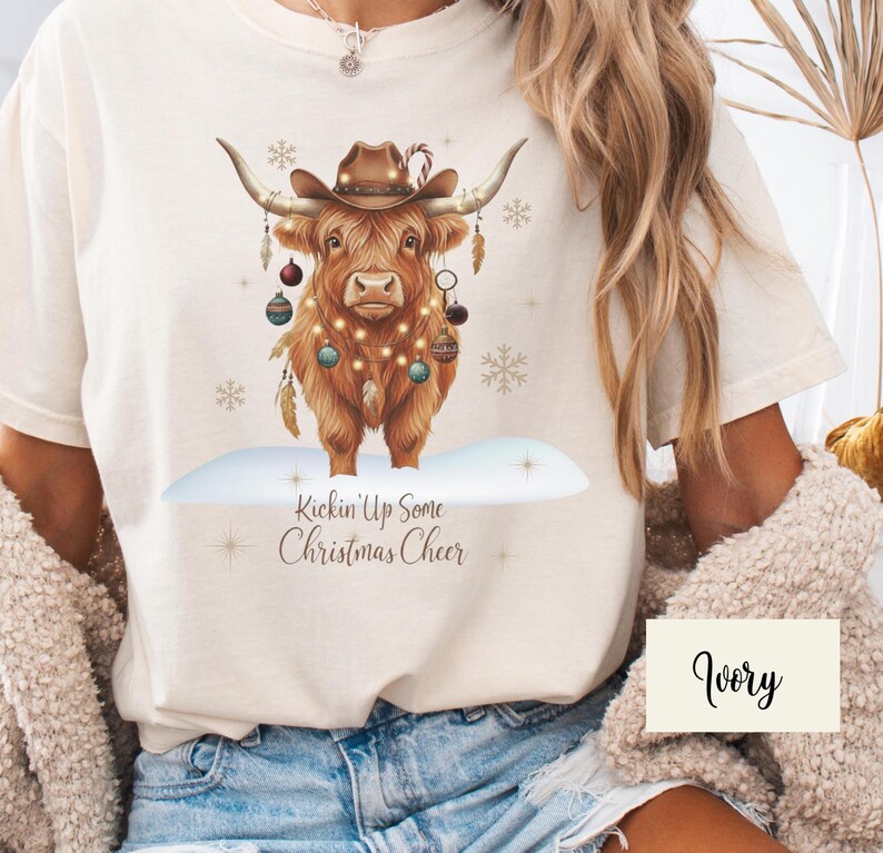 Puede incluir: Camiseta color marfil con una ilustraci&oacute;n festiva de una vaca de las Tierras Altas. La vaca lleva un sombrero de vaquero y est&aacute; adornada con adornos y luces navide&ntilde;as. El texto "Kickin' Up Some Christmas Cheer" est&aacute; debajo.