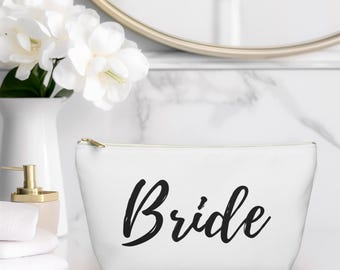 May include: White cosmetic bag with the word "Bride" in black script. The bag has a gold zipper and is sitting on a white surface. A vase of white flowers and a gold-toned soap dispenser are in the background.