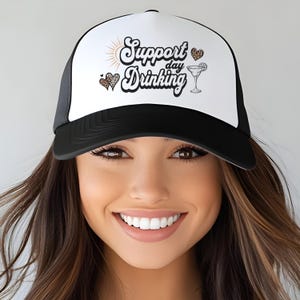 May include: Black and white trucker hat with the text "Support Day Drinking" in a script font. The hat features a white front panel with a sun graphic and leopard print hearts. A cocktail illustration is also on the front.