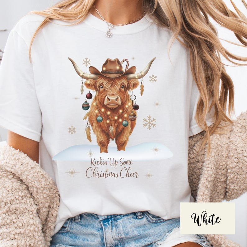 Puede incluir: Camiseta blanca con una vaca de las Tierras Altas marr&oacute;n que lleva un sombrero de vaquero y luces navide&ntilde;as. La vaca est&aacute; decorada con adornos y plumas, de pie sobre un suelo nevado. El texto "Kickin' Up Some Christmas Cheer" est&aacute; debajo.