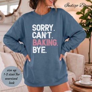 May include: An indigo blue crewneck sweatshirt featuring the white text SORRY. CAN'T. and the pink text BAKING. followed by the white text BYE. The garment has long sleeves and a ribbed collar, cuffs, and hem, with a distressed font style.