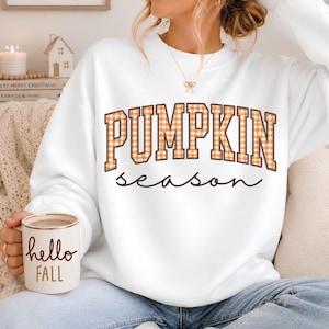 May include: A white long-sleeve sweatshirt featuring the text PUMPKIN SEASON in orange and white gingham block letters with a dark brown outline. A white ceramic mug is held in the foreground with the brown text hello FALL printed on the front.
