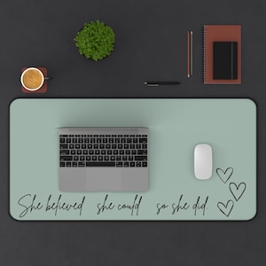 May include: A light green desk mat with the text "She believed she could so she did." written in black cursive. There are three black heart shapes drawn on the right side of the mat. A laptop computer, a white mouse, and a small potted plant are on the mat.