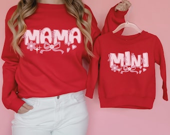 May include: Red sweatshirts, one adult and one child-sized, with matching designs. The adult sweatshirt says "MAMA" in pink bubble letters with floral and bow accents. The child's sweatshirt says "MINI" in a similar style. Both are long-sleeved.
