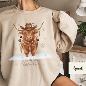 Puede incluir: Sudadera color arena con una ilustraci&oacute;n festiva de una vaca de las Highlands. La vaca lleva un sombrero de vaquero y est&aacute; adornada con adornos y luces navide&ntilde;as. El texto "Kickin' Up Some Christmas Cheer" est&aacute; debajo.