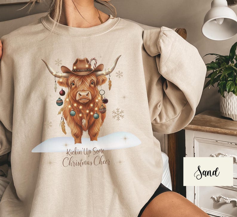 Puede incluir: Sudadera color arena con una ilustraci&oacute;n festiva de una vaca de las Highlands. La vaca lleva un sombrero de vaquero y est&aacute; adornada con adornos y luces navide&ntilde;as. El texto "Kickin' Up Some Christmas Cheer" est&aacute; debajo.