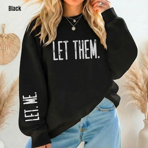 May include: Black sweatshirt with the text "LET THEM." printed in white across the chest. The left sleeve has the text "LET ME." in white. The sweatshirt is paired with blue jeans.