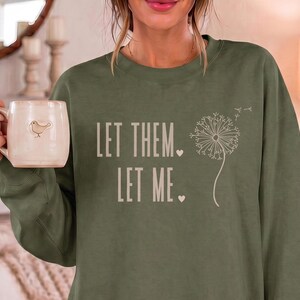 May include: An olive green crewneck sweatshirt featuring the text "LET THEM. LET ME." in a light beige font, accompanied by a graphic of a dandelion seed head and two small hearts. A light-colored ceramic mug with a small bird icon is also visible.