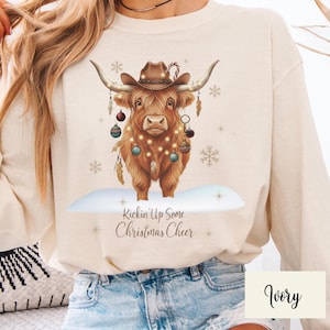 Puede incluir: Sudadera color marfil con una vaca de las Tierras Altas marr&oacute;n que lleva un sombrero de vaquero y adornos navide&ntilde;os. La vaca est&aacute; sobre una superficie nevada, con el texto "Kickin' Up Some Christmas Cheer" debajo.