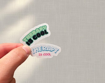 May include: Two stickers with the phrase "THERAPY IS COOL" in green and blue lettering. One sticker has a green border, and the other has a blue border. The stickers are held up against a light gray background.