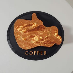 May include: A copper-colored 3D printed mountain range on a black circular base with the word "COPPER" printed below the mountain range.