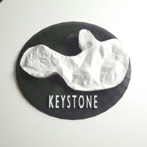 May include: A white 3D printed model of a mountain range on a black circular base with the word "KEYSTONE" printed in white.