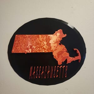 May include: A red and white outline of the state of Massachusetts on a black circular background. The word "MASSACHUSETTS" is written in red below the state outline.