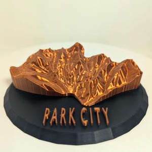 May include: A 3D-printed topographical model of Park City, Utah, in shades of brown and orange. The model sits on a black, circular base with "PARK CITY" in raised lettering.