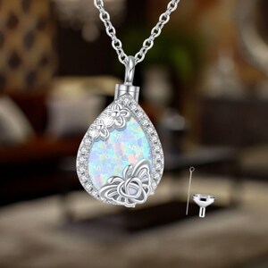 May include: Silver teardrop-shaped pendant necklace with a white opal-like stone and a butterfly design. The pendant is a cremation necklace with a small funnel and tool for filling the pendant.