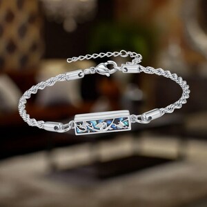 May include: A silver bracelet with a rectangular centerpiece featuring a blue and iridescent inlay with a floral design. The bracelet has a rope-like chain and a lobster clasp closure. The bracelet is approximately 7 inches in length.