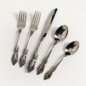 May include: A set of five silver-toned flatware pieces, including two forks, a knife, and two spoons. The flatware has an ornate, decorative design.