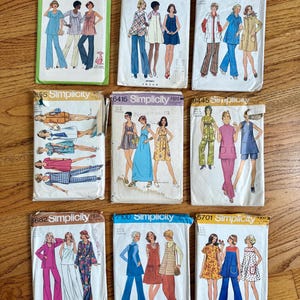 May include: A collection of nine vintage sewing pattern envelopes from Simplicity and Butterick, featuring illustrations of various clothing styles including tunics, dresses, and pantsuits, with printed brand names and pattern numbers.