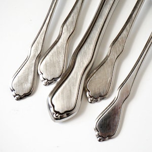 May include: Close-up of several stainless steel spoons arranged diagonally against a white background. The spoons have a classic design with a textured finish and decorative edges. The spoons are likely part of a flatware set.