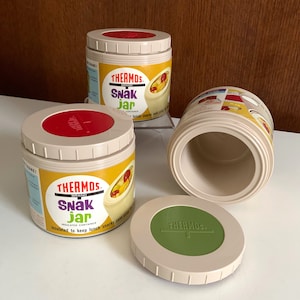 May include: Three vintage Thermos brand insulated snack jars with red and green lids. The jars are beige with yellow and red labels that say "Thermos Brand Snack Jar Insulated Container" and "Insulated to keep lunch snacks cold."
