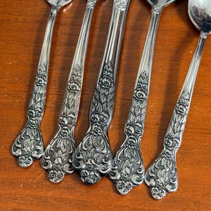 May include: Close-up of silverware with ornate floral detailing. The set includes forks, spoons, and a knife, all with a shiny, silver finish. The handles feature a detailed rose and leaf design, adding a touch of elegance to the dining set.
