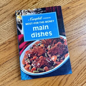 May include: A Campbell's Cookbook titled "MOST-FOR-THE-MONEY main dishes" is displayed. The cover features a photo of a dish with meat, carrots, and green beans in a white oval dish. The background is a wooden surface.