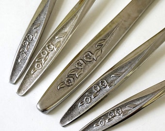 May include: Close-up of several silver-colored utensils with floral detailing. The utensils have a shiny, reflective surface and a decorative pattern of flowers and leaves. The arrangement is on a white background.