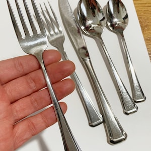 May include: A set of stainless steel silverware including a fork, knife, and spoons. The silverware has a shiny, reflective surface and a geometric design on the handles. The fork is held in a hand, and the set is arranged on a white surface.