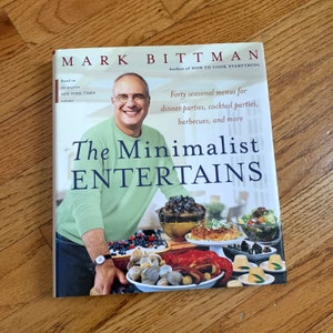 May include: A green book titled "The Minimalist Entertains" by Mark Bittman, author of "How to Cook Everything". The book cover features a photo of the author and a table setting with food.