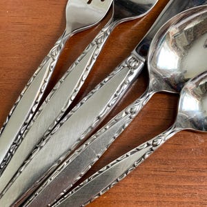 May include: Close-up of stainless steel silverware set on a wooden surface. The set includes forks, spoons, and knives. The silverware has a decorative floral pattern along the handles.