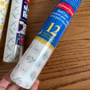 May include: A roll of white shelf liner with a floral pattern. The liner is packaged in a blue and yellow tube with the brand name Rubbermaid and the text "Shelf Liner" in white. The tube also includes the text "10 sq ft (3.3 yd x 12 in) 95m² (3.1 m x 30.5 cm)".
