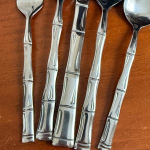 May include: A set of silver-colored flatware with bamboo-style handles. The set includes forks, knives, and spoons, all arranged on a wooden surface. The silverware has a shiny, reflective finish.
