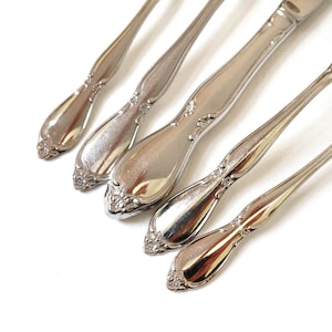 May include: Close-up of five pieces of silverware with a shiny, silver finish. The handles have a decorative floral design at the base and the tip. The silverware is arranged diagonally against a white background.