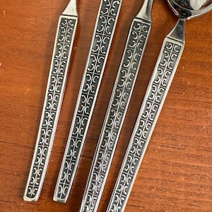 May include: Close-up of four stainless steel spoons with decorative handles. The handles feature a repeating black and silver geometric pattern. The spoons are arranged diagonally on a wooden surface, showcasing their reflective surfaces.