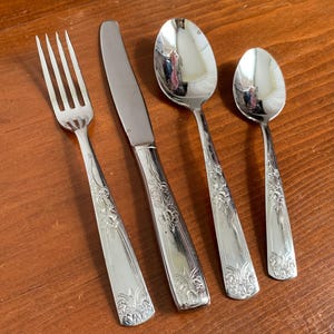 May include: A set of four silver-colored utensils: a fork, a knife, a large spoon, and a small spoon. Each piece has a floral design on the handle. The utensils are arranged on a wooden surface.