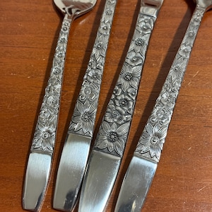 May include: A set of four stainless steel flatware pieces including a fork, two spoons, and a knife. Each handle features a detailed embossed floral pattern with various flower shapes, transitioning into a smooth, plain polished metal base.